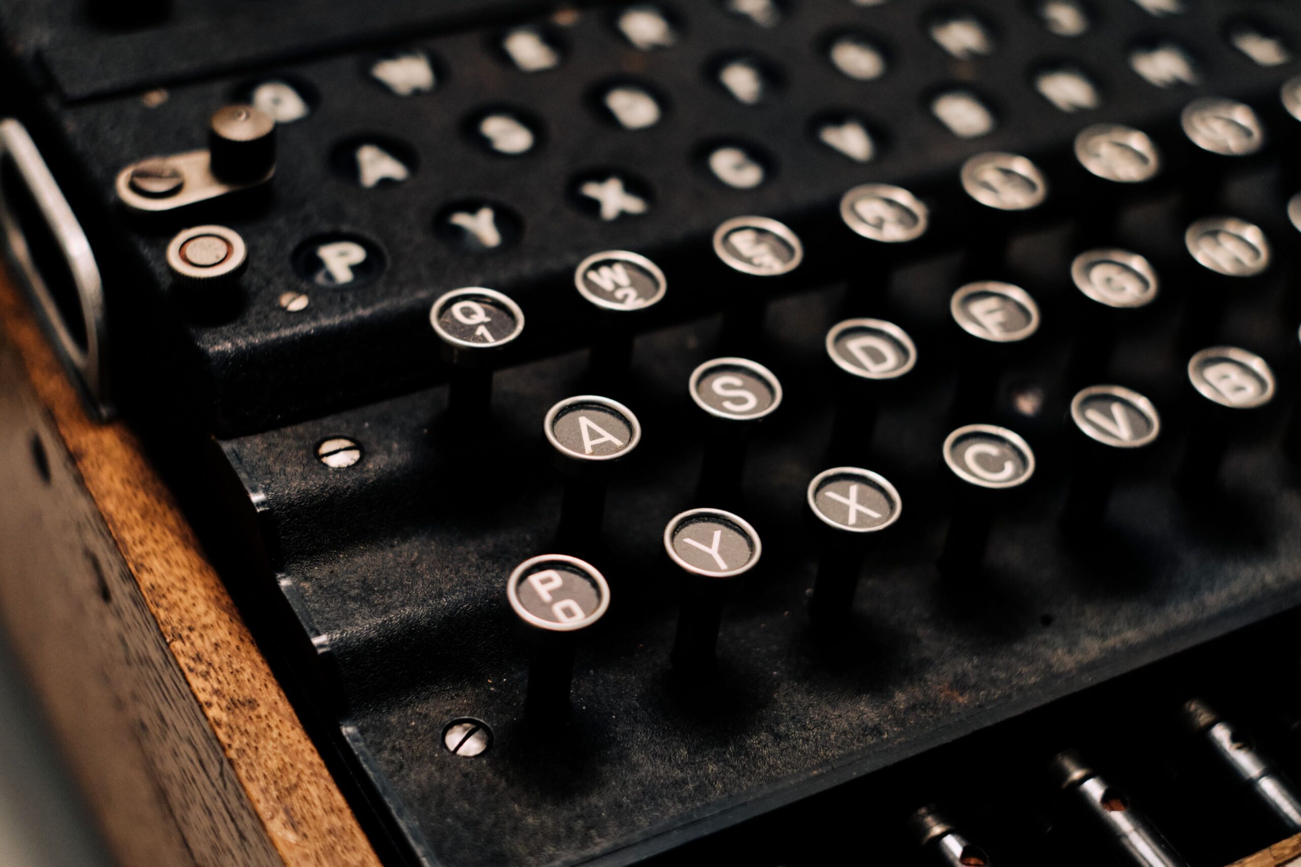 Das Bild zeigt eine Nahaufnahme eines historischen Geräts mit zahlreichen runden Tasten – es handelt sich um die Tastatur einer Enigma-Verschlüsselungsmaschine aus der Zeit des Zweiten Weltkriegs. Die Tasten sind schwarz mit weißen Großbuchstaben in der Mitte, jeweils von einem schmalen silbernen Rand umgeben. Die Buchstaben sind in einer Art QWERTZ-Anordnung zu sehen, ähnlich einer Schreibmaschine, jedoch ohne Zifferntasten oder Sonderzeichen. Oberhalb der Tasten befinden sich kleine runde Sichtfenster mit weiteren Buchstaben, die zur Anzeige der chiffrierten Buchstaben dienten. Die Maschine besteht aus einem schwarzen Metallgehäuse und ist in einem hölzernen Rahmen eingefasst. Die Mechanik darunter ist leicht sichtbar, was dem Gerät ein industriell-technisches Aussehen verleiht. Die Lichtverhältnisse sind warm und erzeugen einen leicht nostalgischen Eindruck.