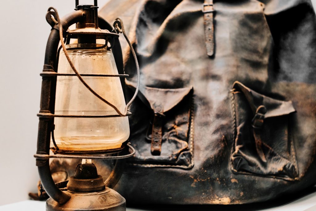 Im Vordergrund des Bildes steht eine alte, metallene Petroleumlampe. Sie hat einen Glaszylinder, der von einem Schutzkäfig aus schwarzem Metall umgeben ist. Das Glas ist etwas verstaubt und am unteren Rand leicht vergilbt. Auf dem Glas ist in französischer Sprache eingeprägt: FABRIQUÉ EN A..., was auf eine französische Herkunft hinweist. Im Hintergrund befindet sich ein großer, abgenutzter Lederrucksack in dunklem Braun. Der Rucksack zeigt deutliche Gebrauchsspuren: Kratzer, Abschürfungen und Verfärbungen sind sichtbar. Zwei große Vordertaschen mit Lederriemen und Metallschnallen befinden sich auf der Vorderseite. Insgesamt vermittelt das Bild eine historische, maritime oder militärische Atmosphäre.